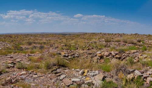 A vast, arid landscape with scattered stone ruins and sparse vegetation under a blue sky with clouds.