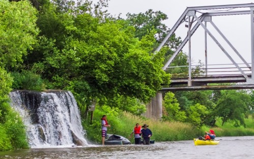 A serene river scene with a small waterfall, a bridge overhead, and people kayaking and gathering by the water's edge.