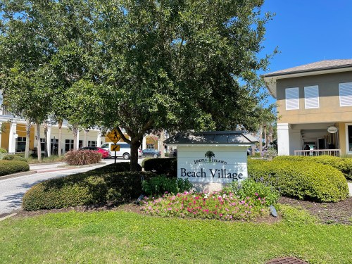 Sign for Beach Village surrounded by greenery and flowers, with buildings in the background under a clear blue sky.