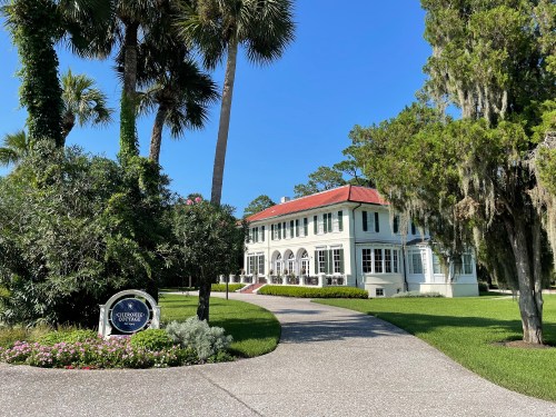 A historic white house with a red roof, surrounded by palm trees and lush greenery, with a sign in front.