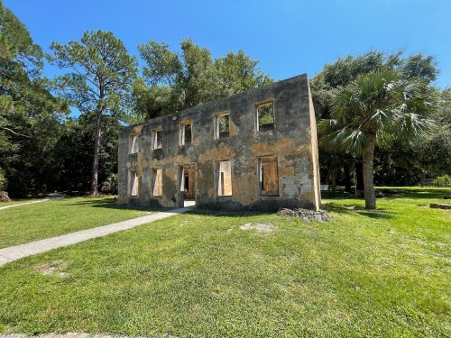 A weathered, two-story building with missing windows, surrounded by green grass and trees under a clear blue sky.