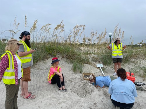 A group of people in safety vests on a beach, observing and working near a sand excavation site.