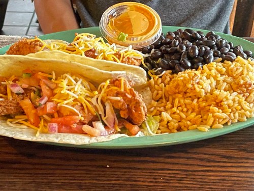 A plate with two tacos filled with meat, cheese, and vegetables, served with rice, black beans, and a small cup of sauce.