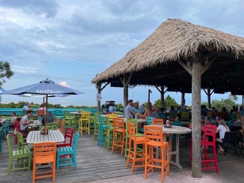 A vibrant outdoor restaurant with colorful chairs, a thatched roof, and diners enjoying the view under a cloudy sky.