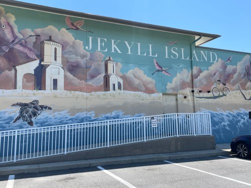 Mural on a building depicting Jekyll Island with birds, a turtle, and a bicycle against a colorful sky.