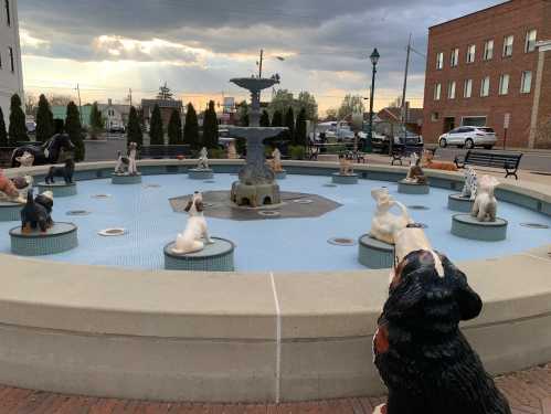 A decorative fountain surrounded by dog sculptures, set against a cloudy sky and buildings in the background.