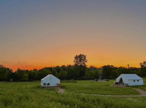 Two tents in a grassy field at sunset, with trees in the background and a colorful sky.