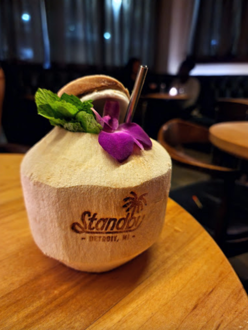 A coconut drink with a metal straw, garnished with a purple flower, sits on a wooden table in a dimly lit restaurant.