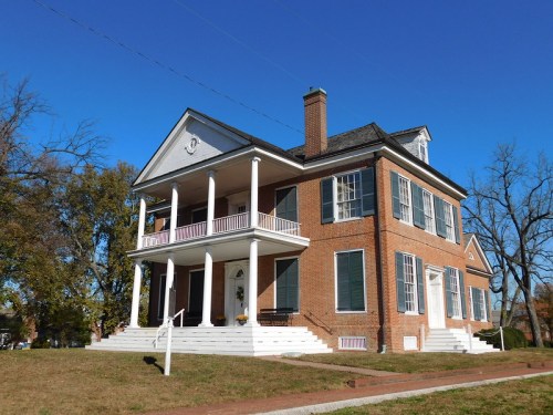 A large brick house with a white porch, green shutters, and a clear blue sky in the background.