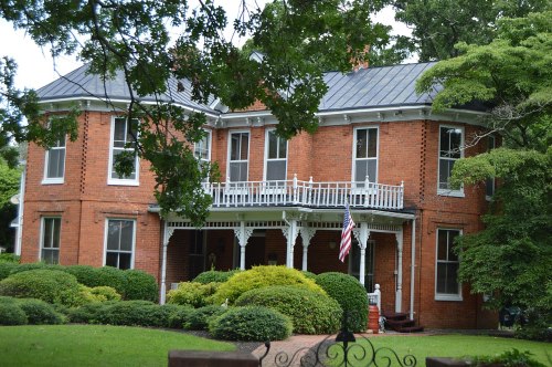 A large red brick house with a porch, surrounded by green bushes and trees, featuring an American flag.