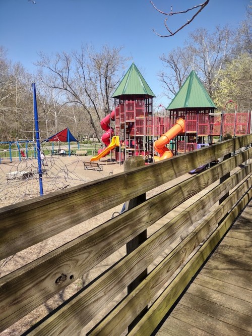 A playground with colorful slides and climbing structures, surrounded by trees and a wooden fence in the foreground.