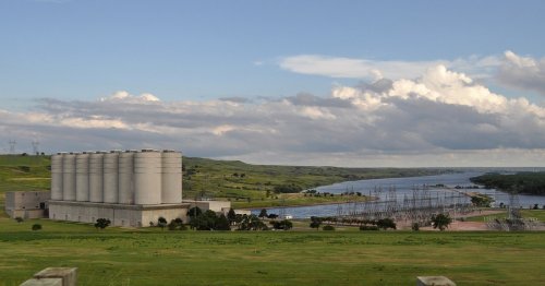 A landscape featuring large silos, a river, and power lines under a cloudy sky, surrounded by green hills.