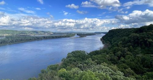 A scenic view of a wide river winding through lush green hills under a partly cloudy sky.