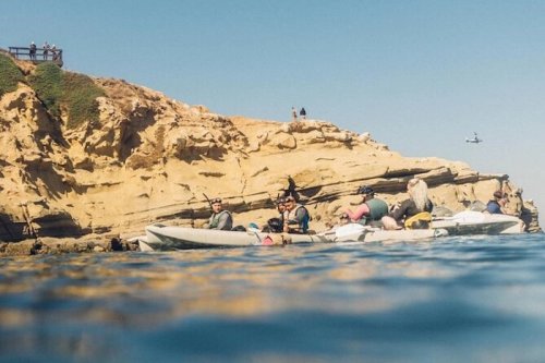 A group of people kayaking near a rocky coastline under a clear blue sky.
