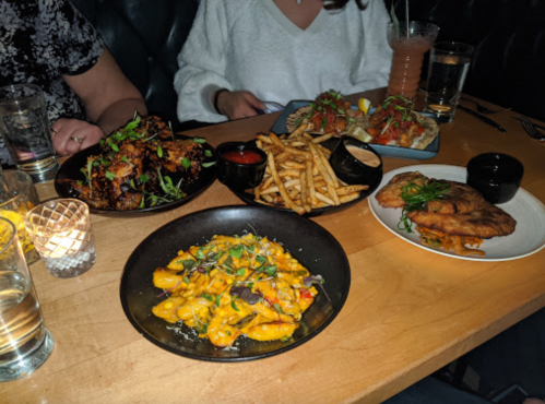 A table with various dishes: wings, tacos, fries, and a creamy pasta, alongside drinks and a candle.