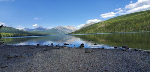 A serene lake surrounded by mountains and lush greenery under a clear blue sky. Pebbly shore in the foreground.