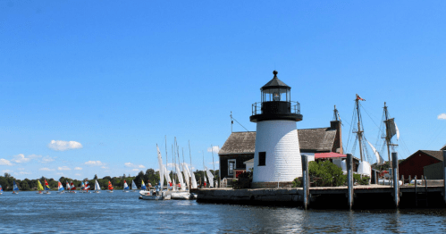 A lighthouse by the water with sailboats in the background under a clear blue sky.