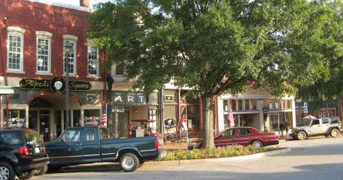 A quaint street scene featuring shops, parked cars, and a large tree in a small town setting.