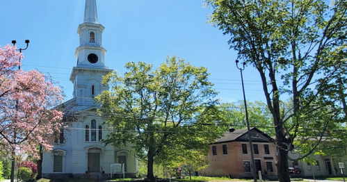 A white church with a tall steeple surrounded by trees and blooming pink flowers on a clear blue sky day.