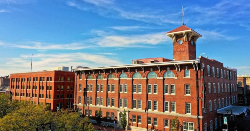 Historic red brick building with a clock tower, surrounded by trees and a clear blue sky.