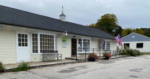 A small, light-colored building with a sign, benches, and American flags, surrounded by flowers and trees.