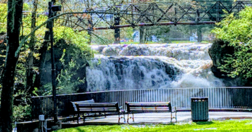 A scenic view of a waterfall with a bridge overhead, surrounded by greenery and benches in a park setting.