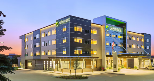 Modern hotel exterior at dusk, featuring large windows and a green logo, with a clear sky in the background.