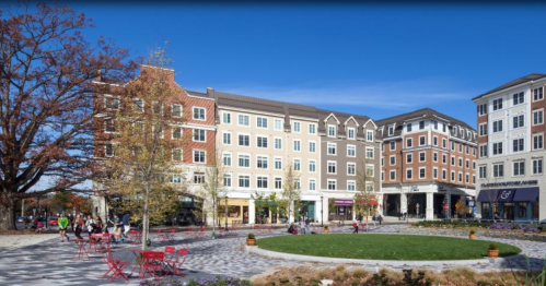 A vibrant plaza with people enjoying the outdoors, surrounded by modern buildings and trees under a clear blue sky.