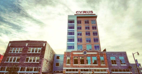A tall building with "CYRUS" on top, surrounded by smaller brick structures under a cloudy sky.