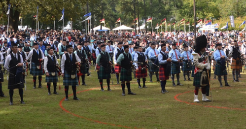 A large group of bagpipers in traditional attire performing outdoors, with a crowd and flags in the background.