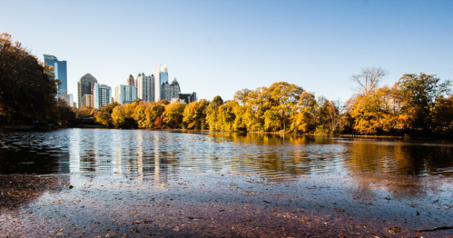 A serene lake surrounded by autumn trees, with a city skyline in the background under a clear blue sky.