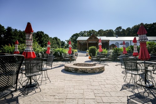 Outdoor seating area with black metal chairs, red umbrellas, and a stone fire pit, surrounded by greenery and a rustic building.