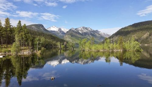 A serene lake surrounded by mountains, with clear blue skies and reflections of trees and peaks on the water's surface.