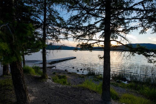 A serene lakeside view at sunset, framed by trees and a wooden dock extending into the calm water.
