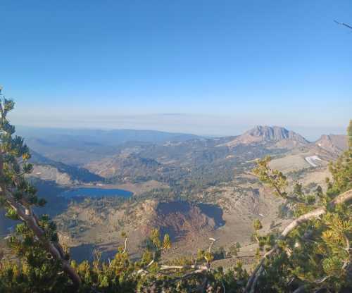 A panoramic view of mountains and valleys under a clear blue sky, with a lake visible in the foreground.