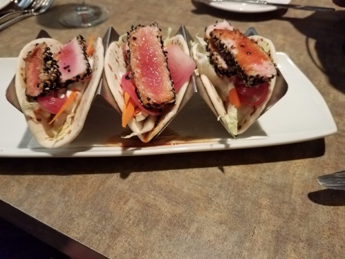 Three tuna tacos with sesame crust, filled with vegetables, served on a white plate.