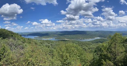 A panoramic view of lush green hills and a winding river under a bright blue sky with fluffy white clouds.