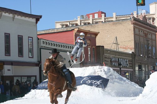 A skier jumps off a snow ramp while a cowboy rides a horse nearby in a snowy street scene.