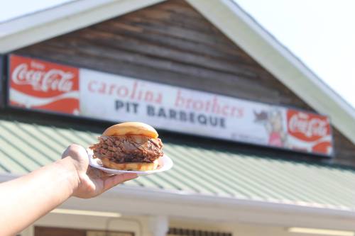 A hand holds a barbecue sandwich in front of a restaurant sign for Carolina Brothers Pit Barbeque.