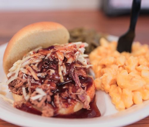 A pulled pork sandwich topped with coleslaw and barbecue sauce, served with macaroni and cheese and green beans.