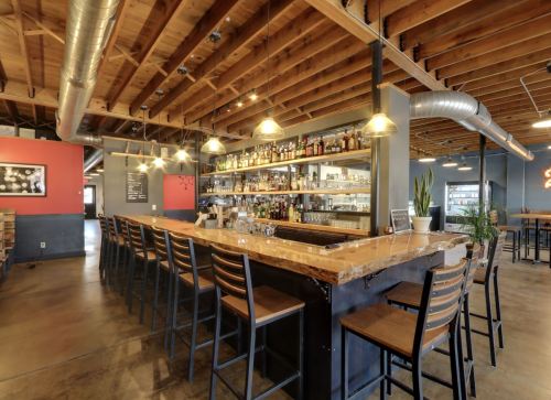 A modern bar with wooden beams, a long counter, shelves of liquor, and minimalist decor in a spacious setting.