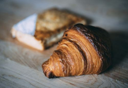 A golden-brown croissant sits in the foreground, with a piece of toasted bread in the background on a wooden surface.