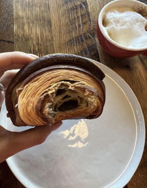 A hand holding a half-eaten croissant on a plate, with a cup of coffee in the background.