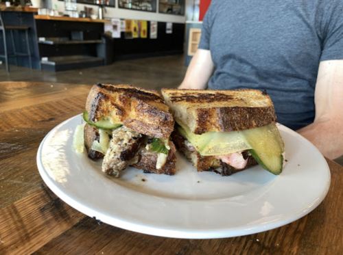 A close-up of a sandwich on a plate, featuring layers of meat, pickles, and greens, with toasted bread.