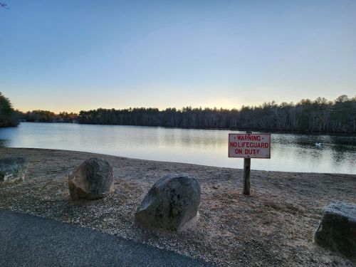 A serene lake at sunset with a sign warning "No Lifeguard on Duty" and large rocks along the shore.