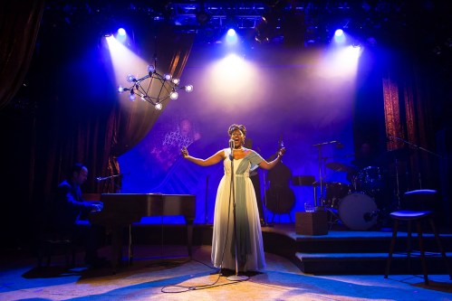 A singer in a flowing dress performs on stage, accompanied by a pianist and a band, under dramatic lighting.