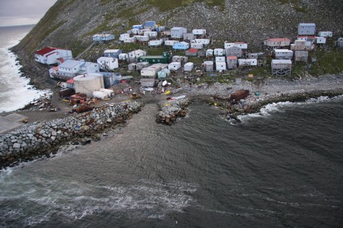 Aerial view of a coastal settlement with colorful buildings on a rocky shore, surrounded by water and hills.