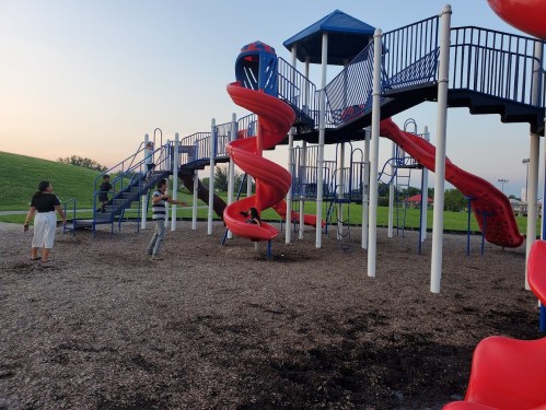 A playground with a red slide and climbing structures, children playing, and green grass in the background at sunset.