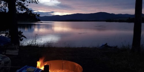 A serene lakeside scene at dusk, featuring a campfire and mountains silhouetted against a colorful sky.