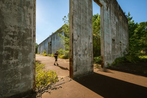 A person walks through the ruins of a concrete structure, surrounded by overgrown vegetation and tall trees.